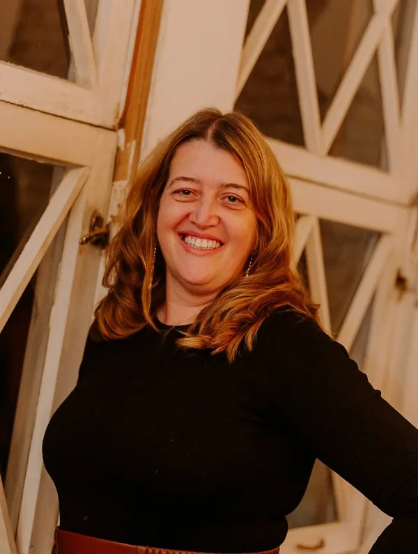 Proprietária e consultora da Triskle Lingeries, sorrindo, de blusa preta, diante de uma janela antiga.